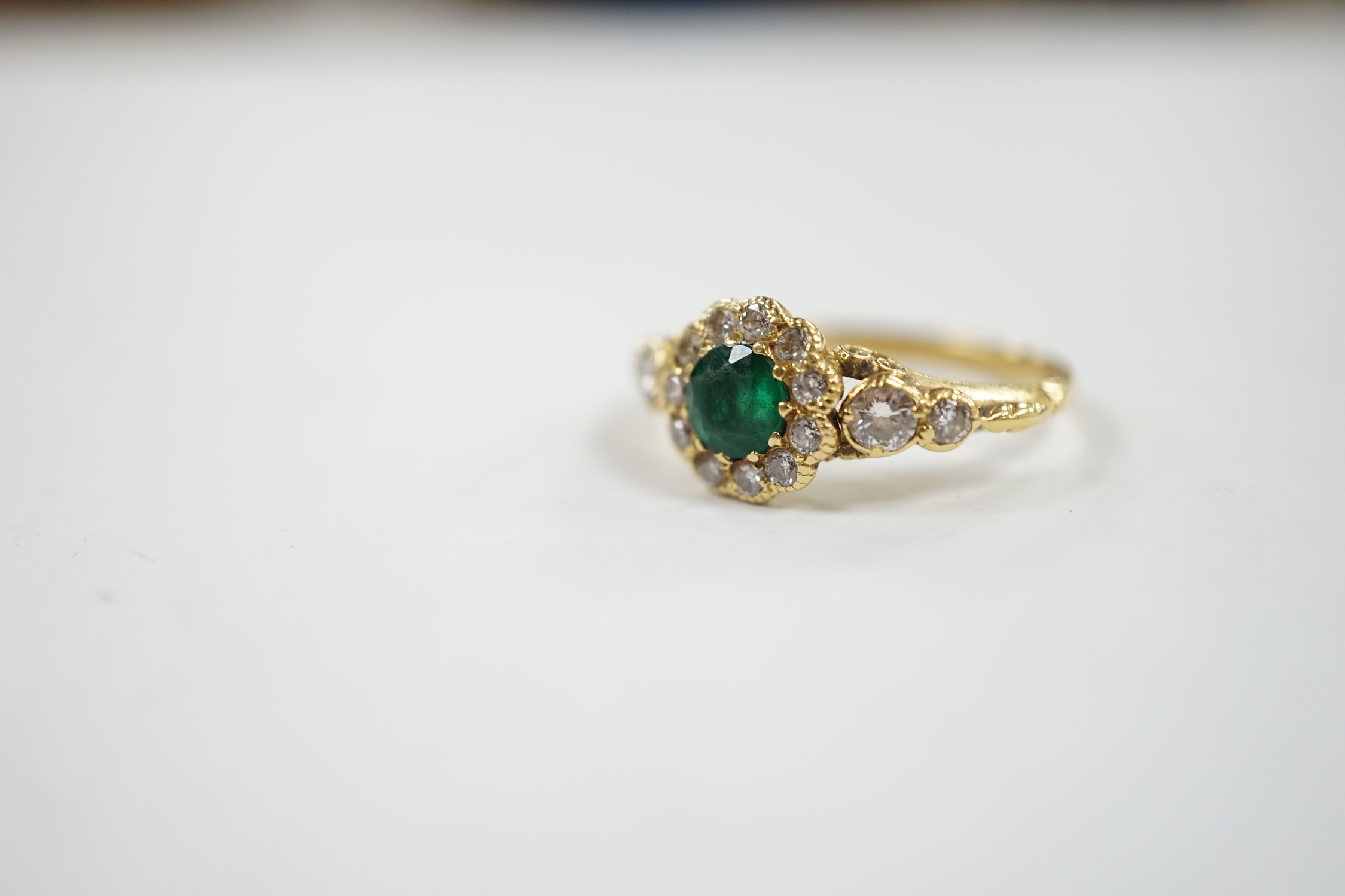 A Victorian style yellow metal emerald and diamond set circular cluster ring, with graduated diamond set shoulders, size N, gross weight 3.7 grams.